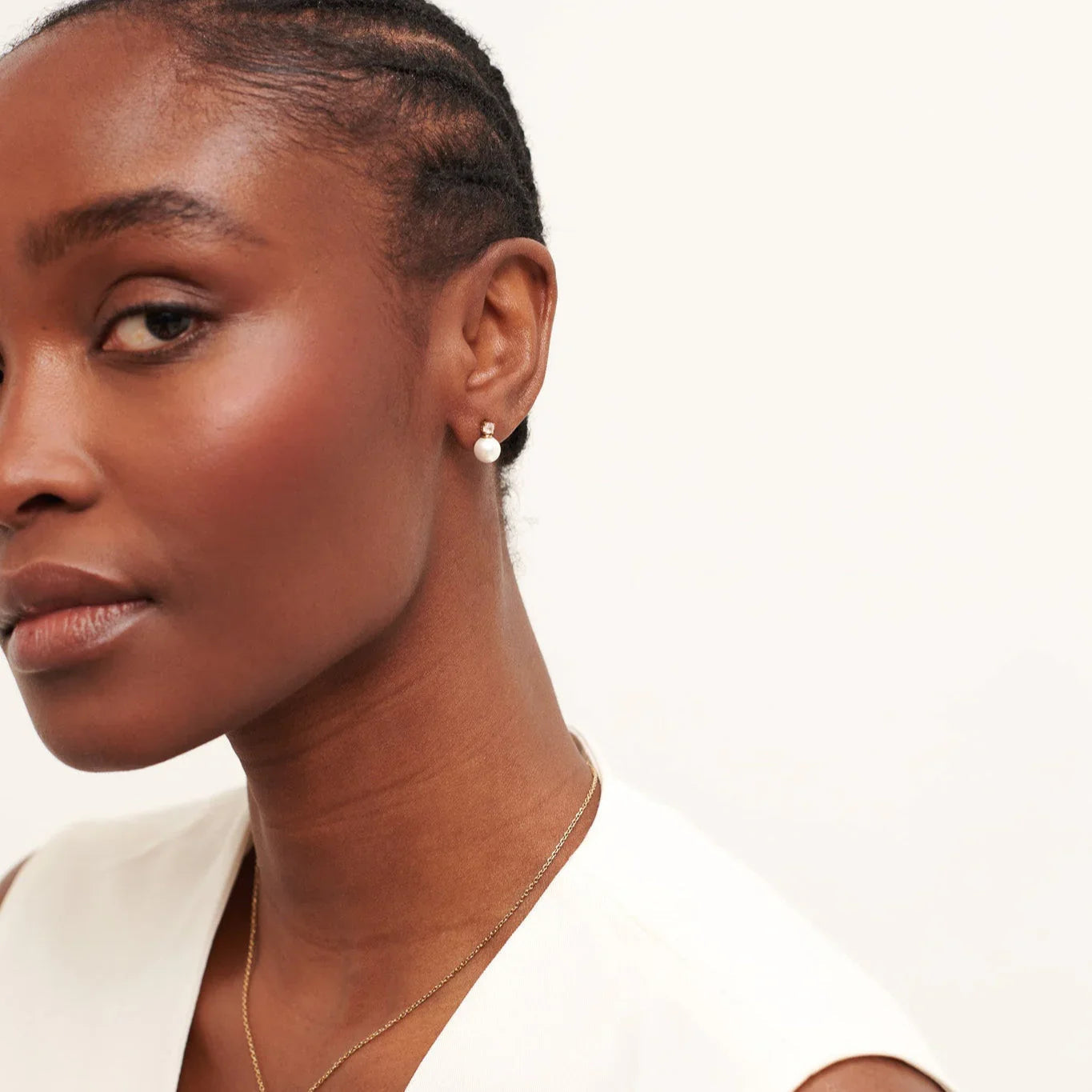 Close-up of a woman with braided hair wearing a white top, pearl stud earrings, and a gold necklace
