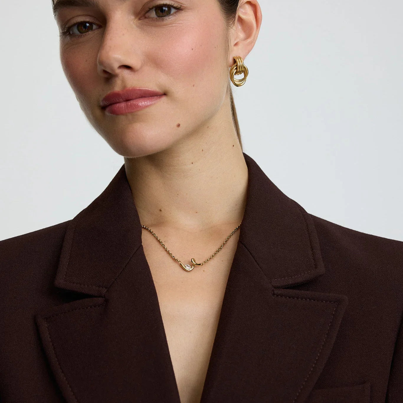 Close-up of woman wearing gold twisted hoop earrings and delicate chain necklace with curved pendant, in brown blazer
