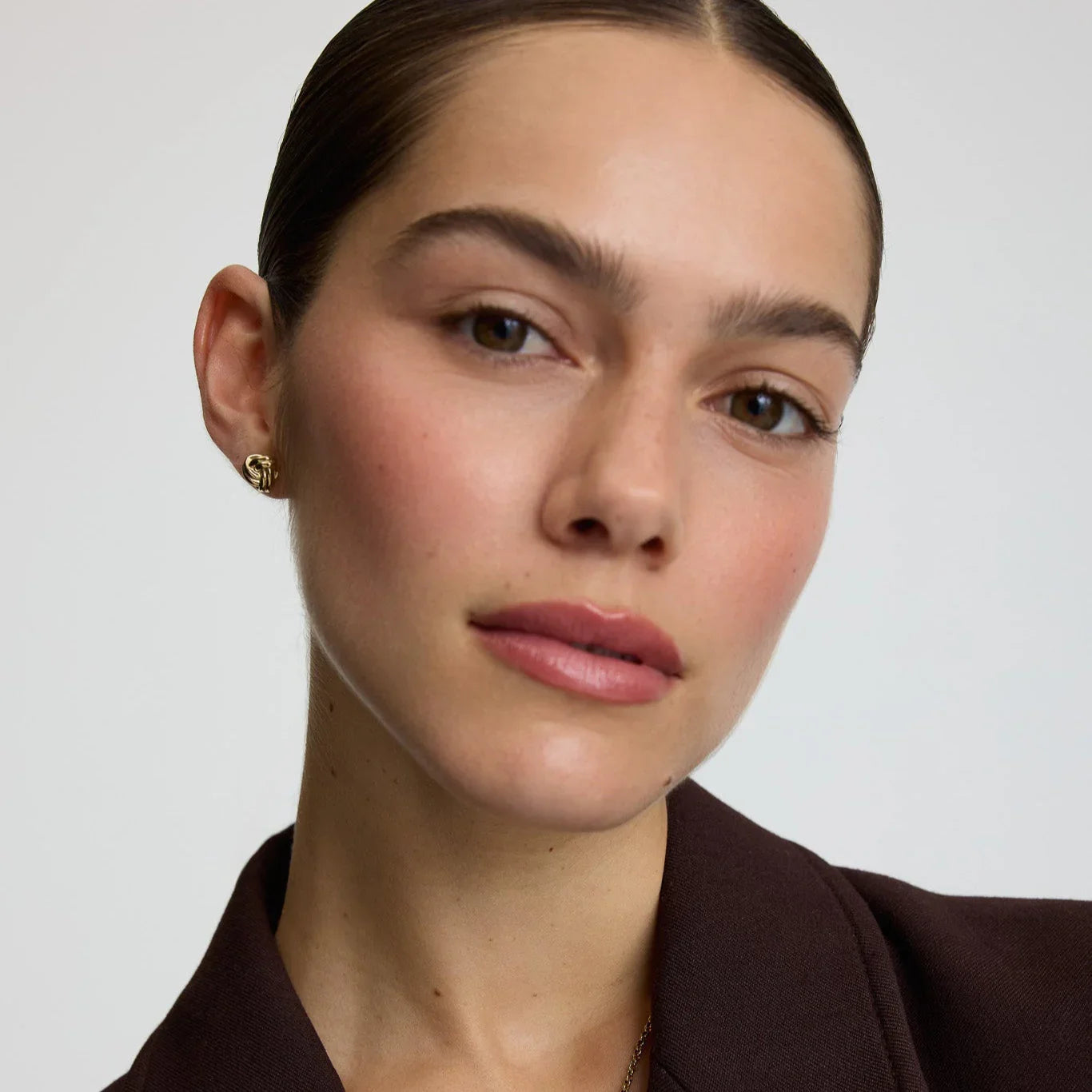 Close-up of woman wearing gold knot stud earrings with sleek hairstyle and brown blazer