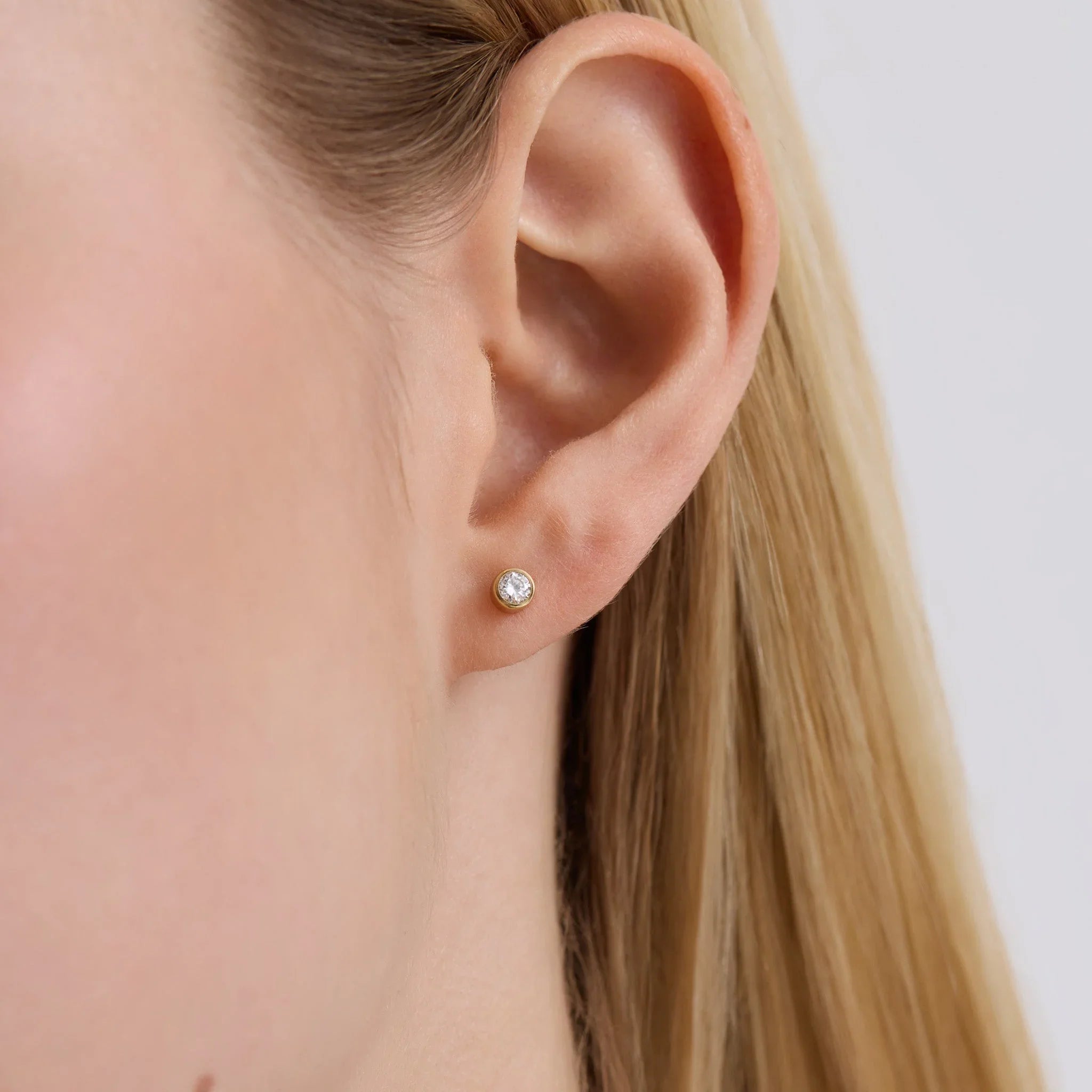 Close-up of a woman's ear wearing a small round diamond bezel stud earring with gold setting