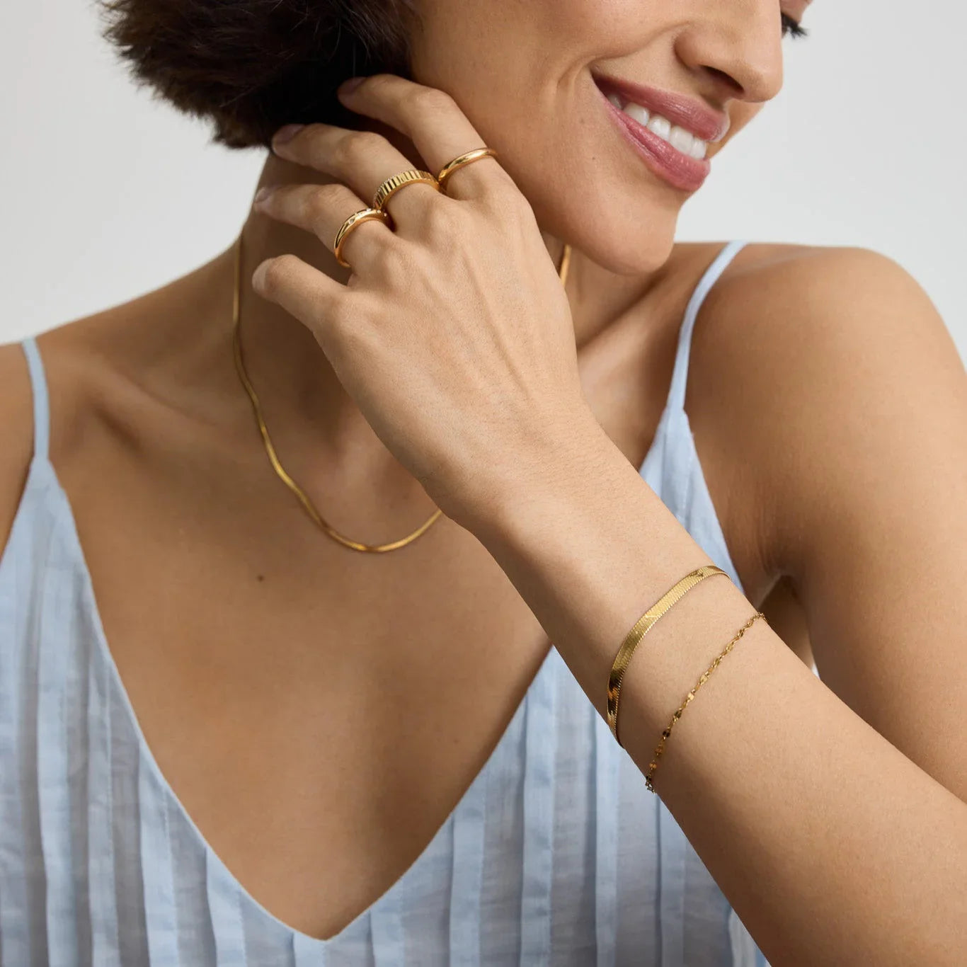 Close-up of woman wearing gold snake chain bracelet, rings, and necklace with light blue striped tank top
